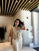 Chaleco sin mangas color beige claro, con escote en V y cierre frontal mediante cordones anudados. Presenta un diseño troquelado con apliques circulares y romboidales en tonos beige y plateado/azul claro.