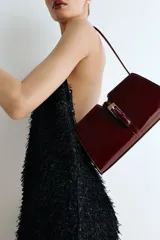 Cartera bandolera color bordó de cuero charolado, con correa fina ajustable y cierre metálico dorado en la parte frontal. Cuenta con un bolsillo interior con cierre y está forrada.