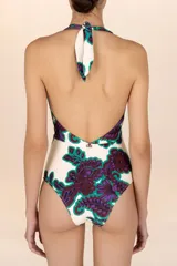Traje de baño entero con estampado floral en tonos morado y verde sobre fondo blanco. Diseño con escote en V pronunciado y tirantes que se atan al cuello.