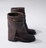 Bota corta de cuero color marrón con suela de goma negra, taco de madera de 3cm y caña de 25cm. Presenta una correa con hebilla y tachas pequeñas alrededor del tobillo.