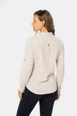Camisa color beige de lino, de manga larga con botones metálicos personalizados y presilla en la manga. Tiene cuello clásico, cierre frontal parcial con botones y bolsillo de parche en el pecho.