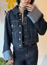 Campera de jean azul oscuro con costuras en contraste color crema, corte cropped, cuello clásico, cierre frontal con botones metálicos plateados, bolsillos de parche en el pecho y mangas largas con puños anchos con dobladillo en color gris. 