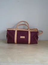 Bolso deportivo grande de lona color borgoña con detalles en cuero color tostado. Tiene doble asa de mano y correa de hombro ajustable y extraíble.