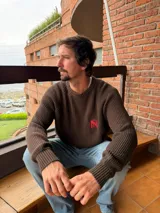 Sweater de punto marrón oscuro con cuello redondo y tejido acanalado grueso en todo el cuerpo y mangas. Presenta un pequeño bordado de iniciales en color rojo en el pecho.