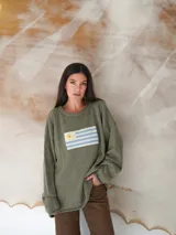 Sweater verde musgo de hilo de algodón tejido con la bandera uruguaya en el frente. Diseño holgado de manga larga.