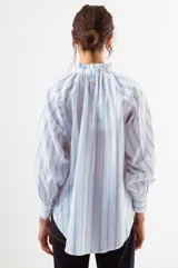 Blusa blanca con finas rayas verticales azules, cuello alto con volado y cierre frontal de botones. Mangas largas abullonadas con puños ajustados.