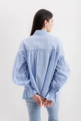 Camisa gris a rayas verticales blancas, de corte amplio, cuello alto con lazo y mangas largas abullonadas con puños elásticos.
