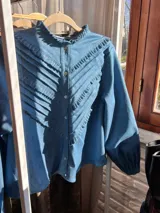 Camisa celeste de jean con cuello mao con volados y volados diagonales en el frente. Tiene mangas largas con puños abotonados y cierre frontal con botones.