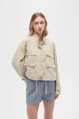 Campera color beige de manga larga, con cuello alto, cierre frontal con cremallera y botones a presión, bolsillos con solapa en el pecho y puños con trabilla ajustable.