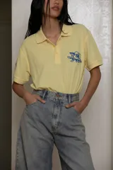 Remera tipo polo color amarillo pálido, de algodón, con cuello clásico y mangas cortas. Presenta un bordado de una rosa azul en el pecho.