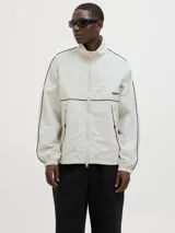 Campera deportiva color crema, de corte recto y cuello alto. Presenta cierre frontal, bolsillos laterales y mangas largas con puños elásticos. Detalle de líneas negras en hombros y mangas, y logo estampado en el pecho.