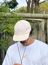 Gorro de visera curva con diseño a rayas verticales en tonos rosado pálido y blanco. La corona tiene un bordado en azul claro con texto. La visera es de color beige claro. Incluye un cordón negro ajustable para colgar del cuello.