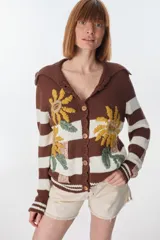 Cardigan tejido de punto color marrón con rayas horizontales blancas y bordado de flores amarillas. Tiene cuello marinero, cierre con botones de madera y corte holgado.