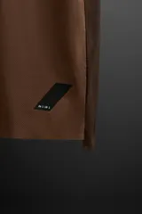 Remera deportiva marrón de tejido técnico, ligero y elástico con estructura perforada. Cuello redondo, manga corta, bajo asimétrico con aberturas laterales, costuras termofijadas y logo estampado.