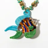 Collar con dije de pez de vidrio multicolor, con detalles en azul, naranja y verde. Cordón trenzado color verde agua con detalles dorados y cuentas doradas.