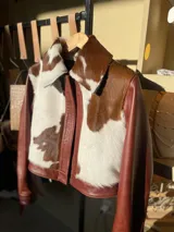 Campera de cuero color marrón rojizo (whisky) con detalles de piel de vaca (marrón y blanco) en el cuello y solapas. Se observa que está abierta, revelando un sweater de punto color crema con botones dorados debajo.