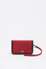Billetera compacta de color rojo con ribetes en contraste de color marrón oscuro. Presenta el logo de Bimba y Lola estampado en blanco en el frente y una correa de muñeca desmontable sujeta con un mosquetón metálico.