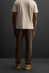 Pantalón deportivo color marrón oscuro, corte regular fit, confeccionado en tejido técnico ligero y elástico. Cuenta con cintura elástica ajustable con cordón, bolsillos frontales con cierre de cremallera, bolsillo trasero con cierre de cremallera oculta en costura y bajo ajustable con elástico.