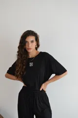 Remera de manga corta color crema, con textura acanalada y cuello redondo. Presenta un pequeño parche bordado con un diseño floral o geométrico en el pecho.