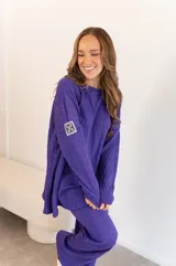 Conjunto de buzo y pantalón de viscosa color violeta con textura. El buzo es de corte holgado con cuello redondo y manga larga con logo en el puño. El pantalón es de corte amplio.