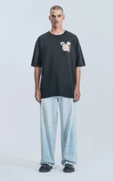 Remera negra de calce oversize, con cuello redondo y un pequeño bordado en el pecho que representa un dim sum (bollo cocido al vapor) en una vaporera de bambú, acompañado de dos platos pequeños y palillos.