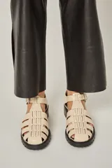 Sandalias de cuero color crema, con diseño de tiras entretejidas y hebilla ajustable en el tobillo.