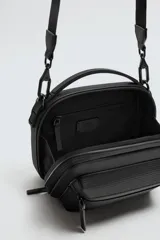 Bandolera negra con acabado engomado y textura acanalada. Tiene un compartimento principal con cierre de cremallera, un bolsillo interior con cierre y un bolsillo frontal con cierre. Incluye asa de mano y asa de hombro ajustable y extraíble.