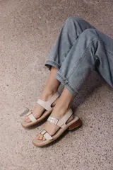 Sandalia plana de cuero color beige claro, con dos tiras anchas ajustables con hebillas metálicas cuadradas de color dorado. El borde de la plantilla está decorado con tachas metálicas doradas.