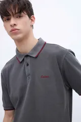 Chomba blanca de algodón con cuello clásico, media cartera de botones, bordado con la palabra 'Uniform' en el pecho y puños con rayas azul marino y rojo.