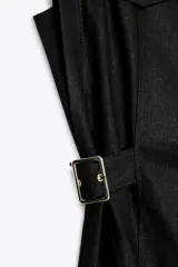 Vestido midi negro, sin mangas, con cuello solapa, escote pico y cierre frontal de botones dorados. Presenta bolsillos falsos con solapas en el pecho y trabillas con hebillas doradas en la cintura.
