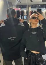 Hoodie negro de corte holgado con capucha y cordón ajustable. Estampado frontal con la leyenda "BE TRENDY EST. 2022" en color rosa.
