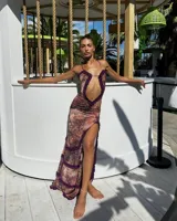 Vestido largo de corte ajustado, con estampado abstracto en tonos rosados y marrones. Presenta un escote pronunciado con detalle de volados en color púrpura, abertura en el abdomen y tajo lateral.