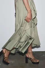 Vestido midi color verde oliva, de cuello redondo y sin mangas. Presenta un cinturón con hebilla metálica y detalle de volantes en la falda.