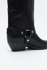 Bota alta de caña recta, color negro, con acabado liso. Presenta punta afilada, taco bajo y estable, y un detalle de arnés desmontable con herrajes metálicos plateados alrededor del tobillo.