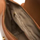 Mochila color habano de material símil cuero (PU) con interior textil rosa viejo. Presenta un asa corta superior, correas traseras regulables y herrajes metálicos dorados. Posee un bolsillo interno con cierre y un bolsillo frontal con solapa y broche metálico.