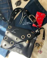 Cartera bandolera de cuero negro con ojalillos metálicos plateados en el frente y correa regulable con hebilla. Tiene un bolsillo exterior y gancho interior para llaves.