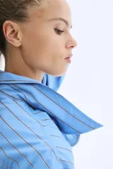 Vestido camisero de corte evasé y holgado, color celeste con finas rayas verticales en negro. Presenta cuello de pico, botonadura frontal y mangas largas con puños abotonados.