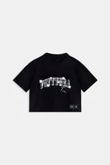 Buzo corto (crop) unisex negro de manga corta y cuello redondo, con estampado frontal en blanco que dice "FUTTTURA TINI" con efecto brillante o desgastado. Incluye una etiqueta rectangular negra con logos en la parte inferior derecha.