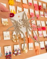 Aretes colgantes grandes con forma de flor estilizada, completamente cubiertos de pedrería brillante color oro. El centro presenta cristales más grandes y facetados.