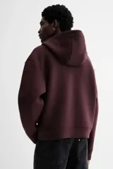 Campera con capucha en tejido efecto neopreno, color borgoña oscuro, con cierre frontal plateado y bolsillos canguro delanteros. Presenta un corte holgado y hombros caídos.