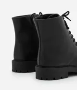 Bota de caña corta para mujer confeccionada en poliuretano (PU) color negro. Presenta punta redonda, suela tipo track con taco mediano de 3,7 cm, cordones frontales con pasadores metálicos y cierre de cremallera lateral.