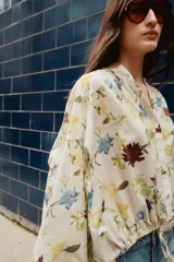 Camisa color crema con estampado de flores en tonos azules, amarillos y borgoña. Tiene cuello redondo con escote en pico, mangas largas con lazos y bajo ajustable con cordones.