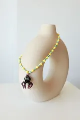Collar de cuentas de plástico verdes y blancas alternadas, con dije de pulpo rojo de vidrio. El dije de pulpo tiene ojos blancos con pupilas negras.