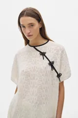 Blusa color crema tejida calada de manga corta con cuello redondo y cierre lateral con lazos negros.