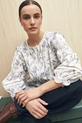 Blusa blanca de algodón bordado con estampado floral en tonos lila y verde, con cuello redondo con encaje, mangas 3/4 con elástico y volados en el ruedo.