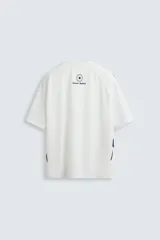 Camiseta blanca de corte holgado con mangas cortas negras. Presenta un logo azul en el pecho izquierdo, un escudo rojo con un caballo en el pecho derecho y la inscripción "Victory Games" en negro en el centro.