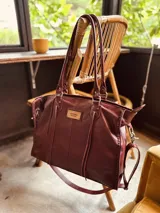 Bolso tipo tote de cuero color borgoña con doble asa de hombro, correa bandolera ajustable y detalles de costuras a la vista.