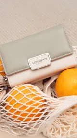Billetera mediana marca Lincoln's confeccionada en cuero sintético con diseño bicolor. Presenta una tapa en tono dorado metalizado con cierre de presilla y cuerpo negro con compartimento de cremallera. Cuenta con múltiples divisiones internas para tarjetas, billetes y monedas, además de una práctica correa de mano.