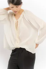 Blusa color crudo de algodón con escote en V y mangas largas abullonadas. Presenta detalles de puntilla y borlas en el escote.