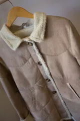 Campera gamulán color beige con cuello y borde de piel sintética, cierre con botones y bolsillos laterales.